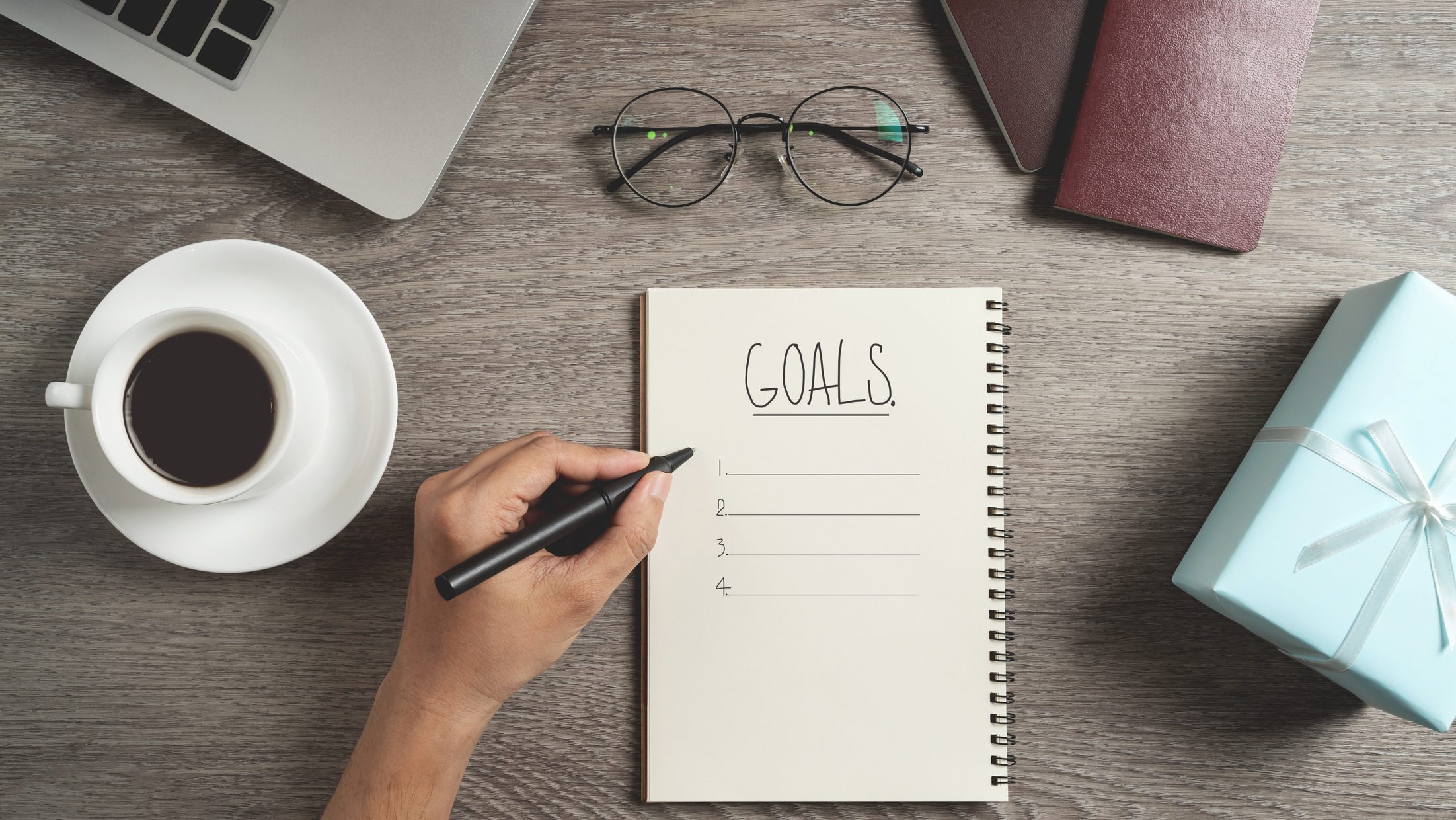 Top view of Man hand is planning and writing to do list and goals in a notebook for New year, Christmas. Gift boxes, cup of coffee, passports, laptop computer and eye glasses on wooden background.